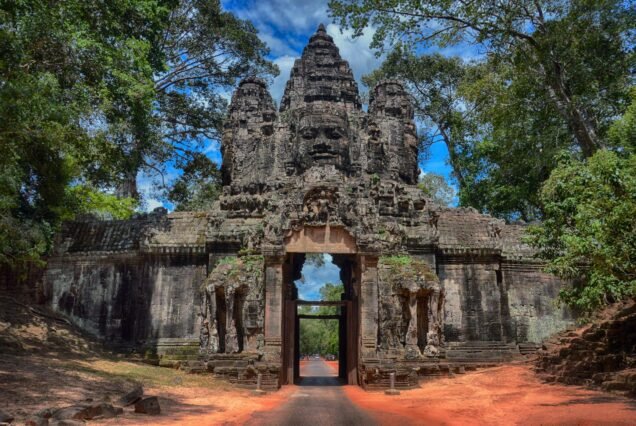 The South Gate of Angkor Thom is a grand 23-meter sandstone entrance featuring four carved faces of the Bodhisattva Avalokiteshvara facing the cardinal directions. Surrounded by dense forest, its central archway serves as an active roadway leading visitors directly into the ancient Khmer city.
